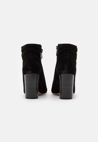 Botines de ante negro con punta redondeada, diseño acolchado en la parte trasera y un tacón grueso y brillante de color negro. Cierre con cremallera lateral.