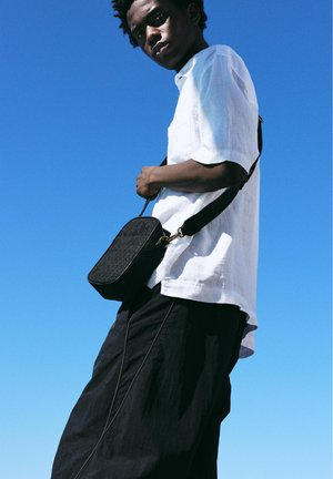 Jonge man die een wit overhemd en zwarte broek draagt, met een zwarte crossbodytas tegen een heldere blauwe lucht als achtergrond.