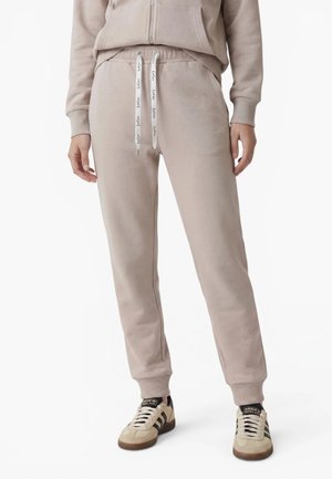 Personne portant un pantalon de jogging beige avec des cordons blancs et des baskets Adidas beige à rayures noires sur fond blanc.