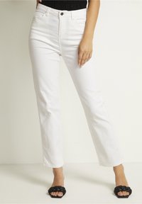 Jeans blancos de pierna recta hechos de denim, con una cintura alta, bolsillos frontales y un cierre clásico de botones. Llevados con sandalias negras de tipo deslizante.