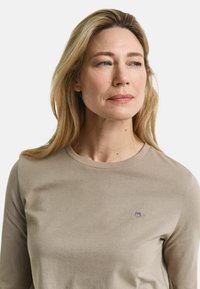 Beige Langarm-T-Shirt aus weicher Baumwolle mit Rundhalsausschnitt und einem kleinen gestickten Logo auf der Brust. Glatte Textur, lässige Passform.