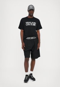 Juodas marškinėlis su "VERSACE JEANS COUTURE" logotipu, juodos šortai, pilkas kepuraitė, baltos kojinės ir juodi sportbačiai. Kasdienis sportinis stilius.