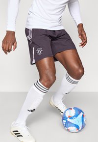 Fekete sportnadrág fehér oldalsávokkal és Manchester United logóval, fehér zoknival és stoplis cipővel, valamint egy kék focilabdával.