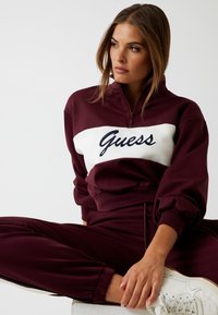Felpa sportiva bordeaux con pannello bianco che presenta "Guess" in scrittura blu navy; tessuto morbido, collo a zip e vestibilità comoda; abbinata a pantaloni coordinati.