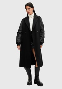 Donna che indossa un lungo cappotto nero con maniche in raso, un maglione bianco a collo alto, una gonna nera e stivali neri al ginocchio, in piedi davanti a uno sfondo semplice.