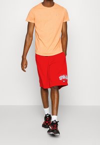 Oranje t-shirt met korte mouwen, gecombineerd met rode sportshorts met een witte "USA" opdruk. Zwarte sportschoenen met rode accenten en witte sokken.