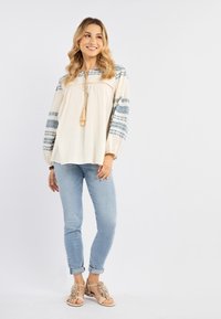 Top leggero di crema con maniche blu a fantasie, abbinato a jeans chiari e sandali piatti beige con dettagli testurizzati.