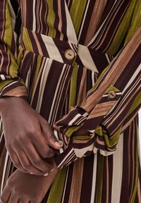 Prążkowana sukienka z pionowymi liniami w odcieniach brązu, oliwkowej zieleni i kremu. Materiał jest miękki z guzikami na mankietach i w talii.