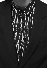 Giacca nera con una finitura testurizzata, indossata su una camicia a righe astratte in bianco e nero con dettagli a bottoni.