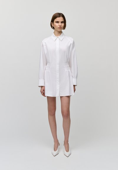 Robe blanche à col pointu, à manches longues, taille froncée et courte, portée avec des talons blancs pointus.