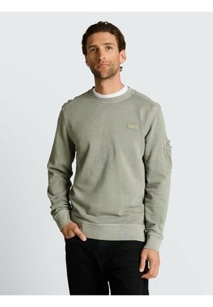 Mężczyzna w jasnzie zielonej bluzie z dekoltem typu crewneck z kieszenią na rękawie i czarnych spodniach, stojący na tle gładkiego jasnego tła.