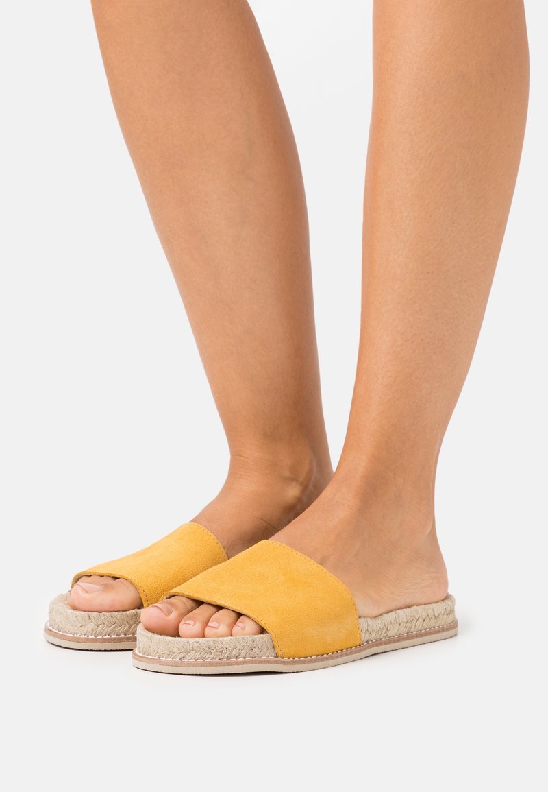 Pieds portant des sandales mules en daim jaune avec semelles plateformes en jute tissé sur fond gris clair.