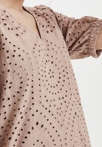 Světle růžová výšivková tunika s výstřihem do V, která má ozdobné otvorové vzory a nabírané rukávy. Hladký povrch s dekorativními švy.