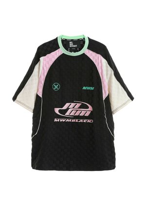 Joven de pie con camiseta deportiva grande en negro, rosa y blanco, pantalones cortos negros, calcetines negros y zapatillas negras gruesas sobre fondo blanco.