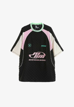 Joven de pie con camiseta deportiva grande en negro, rosa y blanco, pantalones cortos negros, calcetines negros y zapatillas negras gruesas sobre fondo blanco.