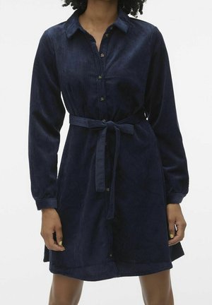 Kvinde iført en marineblå kordfløjls skjortekjole med lange ærmer, knapper foran, krave og bindebælte i taljen.