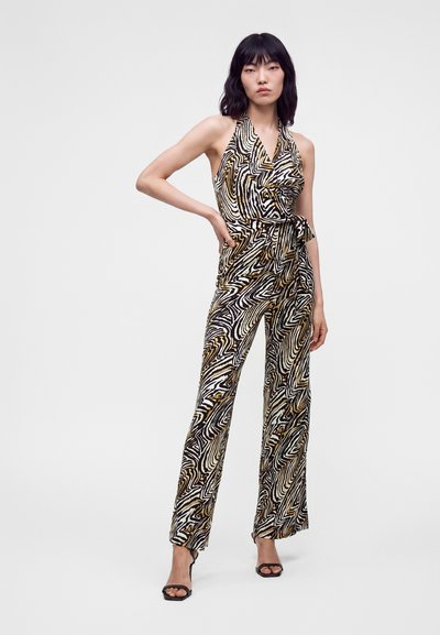 Femme debout, main sur la hanche, portant une combinaison sans manches à motif abstrait noir, blanc et marron et des sandales noires à talons hauts.