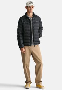 Sort pufferjakke med vandret quiltet mønster, lynlås lukning, båret over en grå skjorte, parret med beige bukser og gule sneakers.