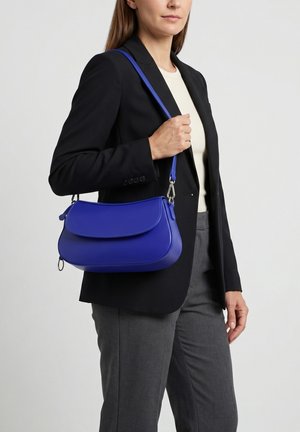 Frau trägt schwarzen Blazer und graue Hose, hält eine leuchtend blaue Schultertasche mit Klappe und Reißverschlussdetail.