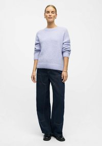 Jasnofioletowy dziergany sweter z prążkowanymi mankietami i ściągaczem, zestawiony z ciemnoniebieskimi dżinsami o szerokich nogawkach z widocznym przeszyciem i kieszeniami. Czarne obuwie.