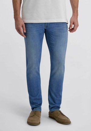 Homme portant un jean bleu délavé, des chaussures beiges à lacets et une chemise blanche, debout devant un fond uni.