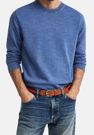 Mann in blauem Langarmpulli, in blaue Jeans gesteckt, mit einem braunen gestreiften Stoffgürtel, Hände in den Taschen.
