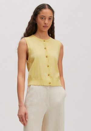 Frau mit langen lockigen Haaren, die ein ärmelloses gelbes, geknöpftes Oberteil und hoch taillierte strukturierte weiße Hose trägt, steht mit einer Hand in der Tasche.