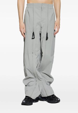 Pantaloni larghi grigio chiaro con due lunghe zip verticali che terminano con tiretti neri, indossati con scarpe da ginnastica nere su una persona in piedi.