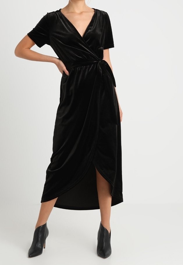 Femme portant une robe portefeuille en velours noir à manches courtes et des bottines, debout avec une main sur la hanche.