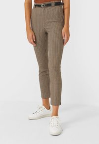 Pantalon ajusté à motif pied-de-poule marron et noir taille haute, associé à des baskets blanches. Il comporte une ceinture noire avec une boucle argentée.