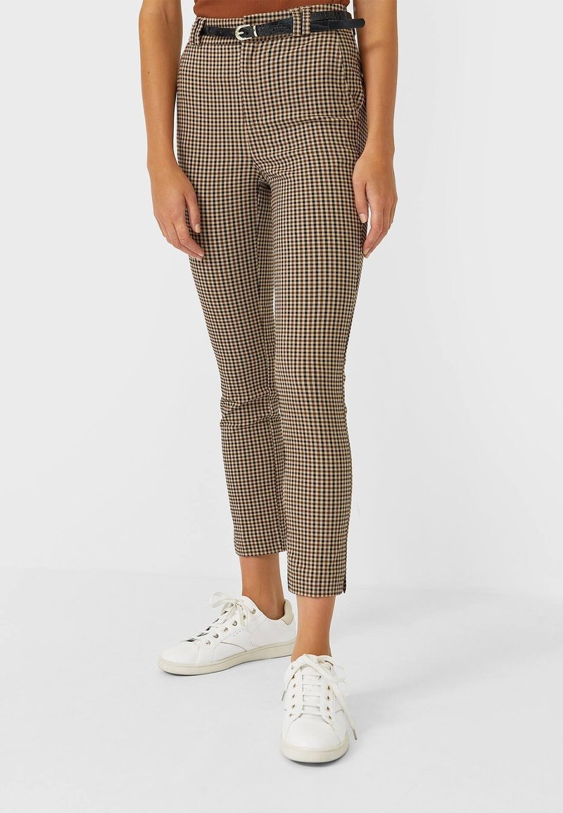 Pantalon ajusté à motif pied-de-poule marron et noir taille haute, associé à des baskets blanches. Il comporte une ceinture noire avec une boucle argentée.