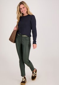 Donkerblauwe langemouwen shirt, groene faux-lederen broek met trekkoord in de taille, bruine tas en bruine sneakers met witte accenten.