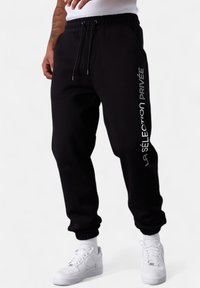 Lower body of person wearing black jogger pants with "LA SÉLECTION PRIVÉE" text on side and white sneakers on a white background.