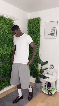 Mann in weißem T-Shirt, grauen Shorts, weißen Socken und dunklen Mokassins steht auf einem grauen Teppich mit grünen Pflanzen und einer Nähmaschine in der Nähe.