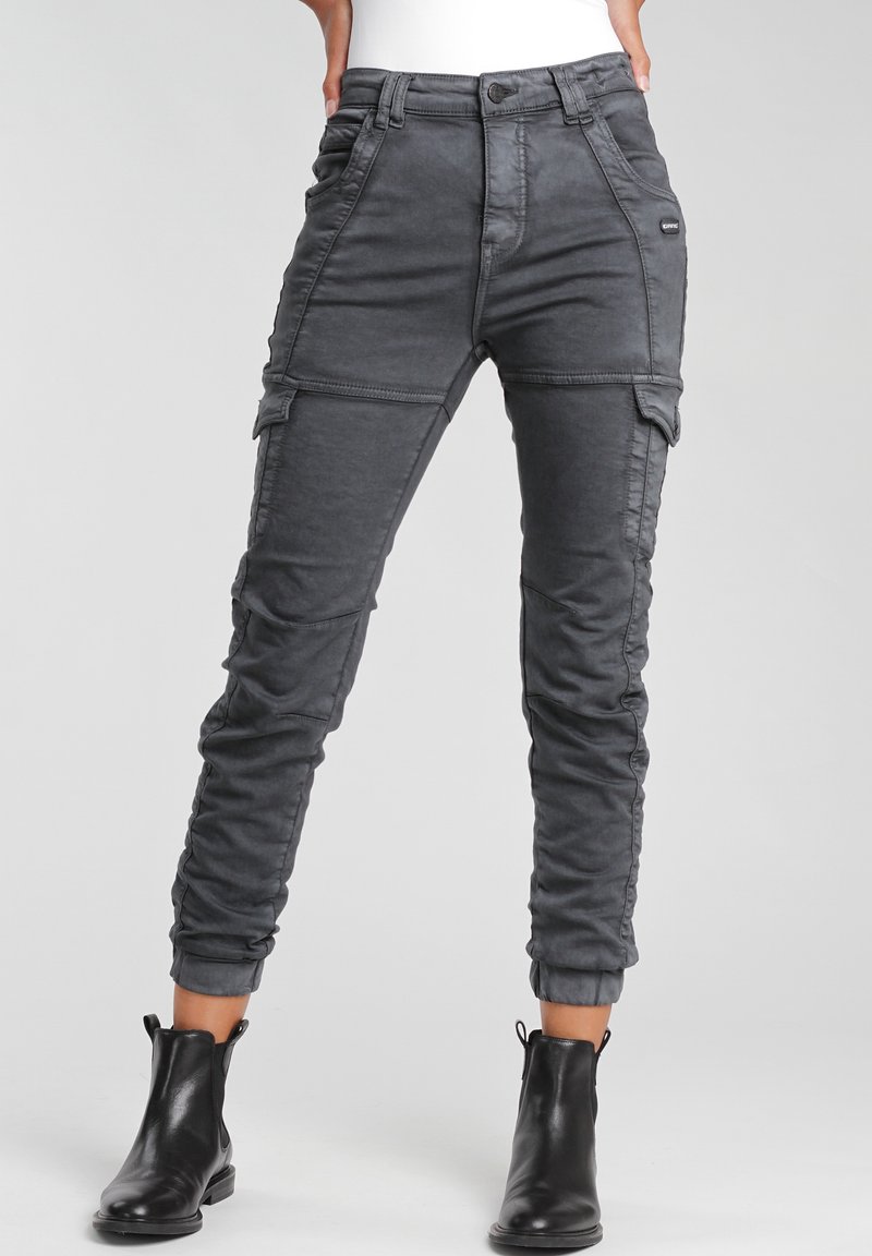 Mujer vestida con pantalones cargo gris oscuro con bolsillos laterales y botas negras hasta el tobillo, de pie frente a un fondo claro y liso.