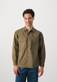 Olive green, long-sleeve shirt made of lightweight fabric with two front pockets and snap-button closure, paired with blue pants.