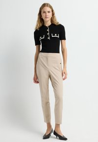 Zwarte geribbelde top met korte mouwen, witte randen en gouden knopen, gecombineerd met beige op maat gemaakte broek en zwarte puntige hakken.
