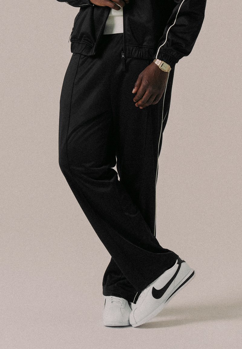 Černé sportovní kalhoty s bílými bočními pruhy, vyrobené z hladkého materiálu, v kombinaci s bílými teniskami s černým logem Nike.