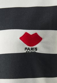 Pruhované bavlněné tričko se horizontálními námořnickými modrými a bílými pruhy, s červeným vyšitým logem rtů a textem "PARIS STUDIO" pod ním.