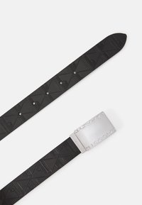 Cinturón de goma negro con un patrón geométrico texturizado y hebilla de metal plateado con el nombre de la marca grabado.