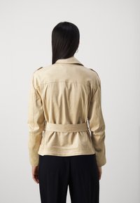 Femme avec de longs cheveux bruns portant une veste beige ceinturée et un pantalon noir, se tenant de dos face à l'observateur contre un fond uni.