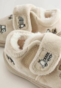 Chaussons pour bébé en beige doux avec doublure en peluche, ornés de camions brodés gris et blancs, et de sangles réglables à boucle auto-agrippante pour un enfilage facile.