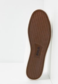 Biały tenis ze strukturą na gumowej podeszwie, z brązowym, wzorzystym spodem i czarnym logo „POLO” po boku podeszwy.