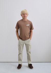 Niño rubio de pie con las manos en los bolsillos, vistiendo una camiseta gráfica marrón, pantalones cargo beiges y zapatillas marrones frente a un fondo liso.