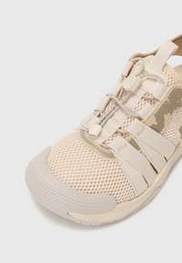 Light beige athletic shoe made of mesh material with elastic straps and a rubber sole. Features a textured surface and minimalistic design.