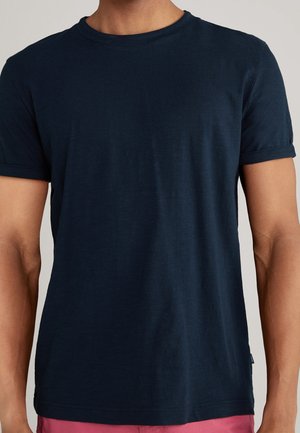 Homme portant un t-shirt à manches courtes bleu marine uni et un short rose, montré du cou à la taille sur un fond uni.