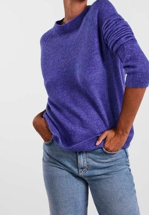 Personne portant un pull violet à manches longues légèrement rentré dans un jean en denim bleu clair, les mains dans les poches, sur un fond uni.