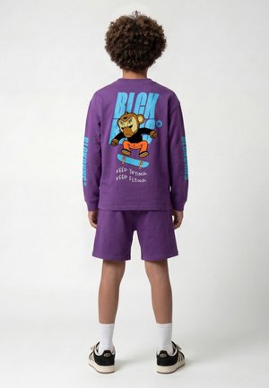 Kind met een paarse lange mouwen shirt en korte broek, met een cartoon aap op een skateboard en de tekst "Keep trying, keep flying" op de achterkant van het shirt.