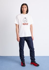 Witte katoenen t-shirt met zwarte grafische print van handen en de tekst "FAIRPLAY"; gecombineerd met donkerblauwe spijkerbroek en rode sneakers.