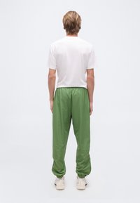 Νεαρός/ή στέκε ται με την πλάτη γυρισμένη, φοράει απλό λευκό κοντομάνικο μπλουζάκι, χαλαρά πράσινα παντελόνια και άσπρα αθλητικά παπούτσια μπροστά σε λευκό φόντο.